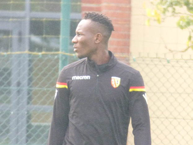 Ein Mann in einer schwarzen Jacke steht auf einem Fußballfeld, umgeben von einem Zaun und einem Baum, und sieht konzentriert während eines Trainingssitzungs.