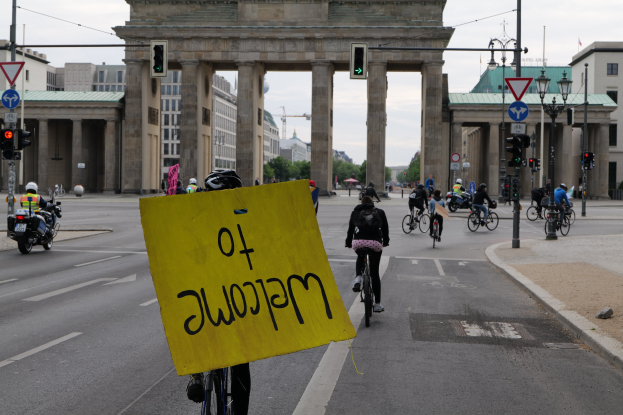 Eine Gruppe von Menschen mit Helmen fährt auf Fahrrädern eine Straße vor dem Brandenburger Tor in Berlin, Deutschland, entlang, wobei eine Person ein gelbes Schild hält, Lichtmasten, Verkehrsampeln, Gebäude, Bäume und einen klaren blauen Himmel im Hintergrund zu sehen sind.