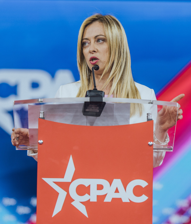 Frau in weißem Kleid, die an einem Rednerpult mit Mikrofon spricht, wahrscheinlich eine Politikwissenschaftlerin, mit unscharfem Hintergrund.