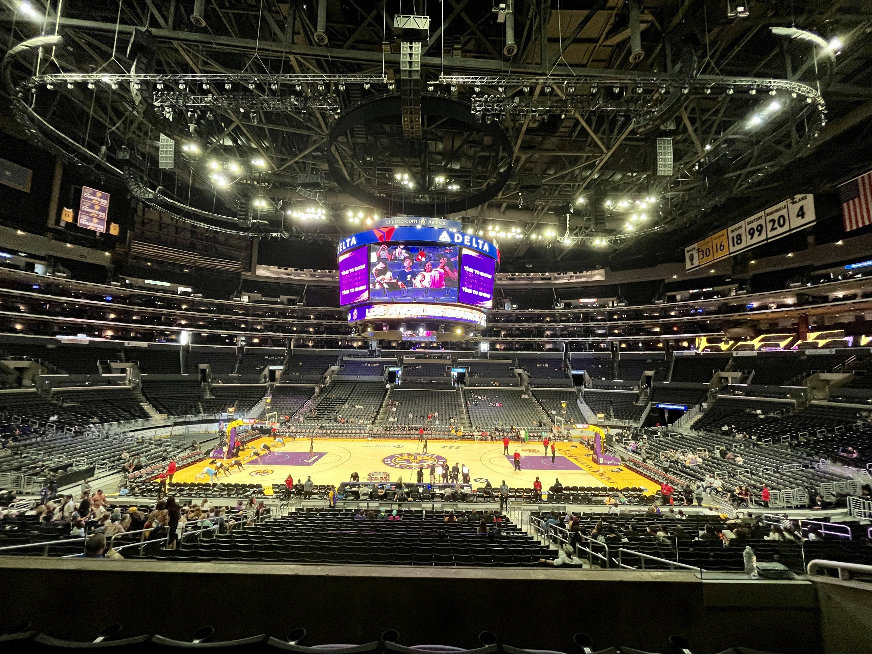 Eine große Indoor-Arena, das Staples Center, voller Zuschauer, die ein Basketballspiel anschauen, mit Sitzbereichen, stehenden Menschen, einem prominenten Bildschirm, einer Flagge, Anzeigetafeln und Deckenbeleuchtung.