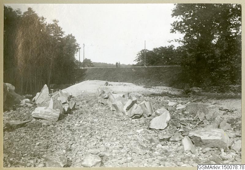 Schwarz-weiß-Foto einer Schotterstraße mit verstreuten Steinen, Bäumen, Strommasten, einer Wand und Himmel, mit Text, der verschiedene Routen am Boden auflistet.