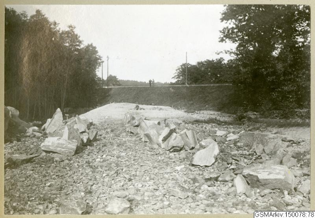 Schwarz-weiß-Foto einer Schotterstraße mit verstreuten Steinen, Bäumen, Strommasten, einer Wand und Himmel, mit Text, der verschiedene Routen am Boden auflistet.