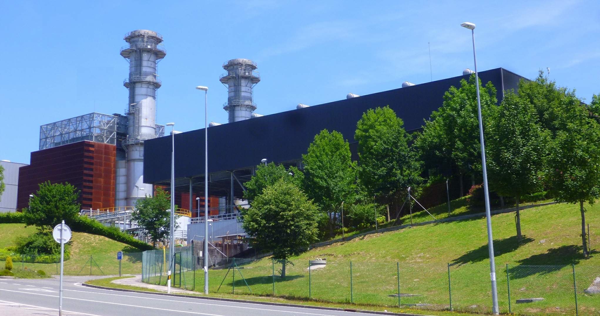 Großes Industriegebäude mit vielen Fenstern, als Kraftwerk identifiziert, umgeben von Straßeninfrastruktur, Fahrzeugen, Grünflächen und einem bewölkten Himmel.