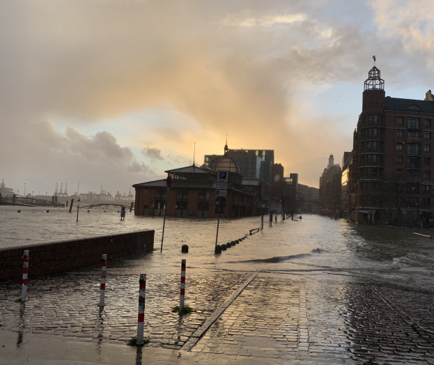 Uberflutete Straße in Hamburg, Deutschland mit Wasser auf der Straße, Masten, Schildern, Gebäuden und einer Brücke unter einem bewölktem Himmel.