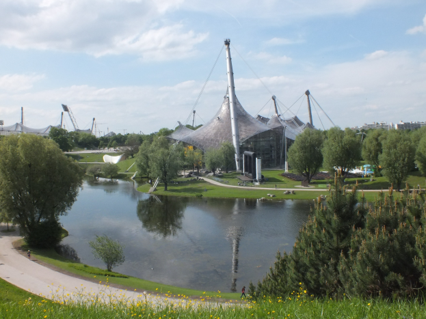 Olympiastadion in London, England, umgeben von Grünflächen, einer Straße, einem Gewässer im Vordergrund und Gebäuden im Hintergrund unter einem bewölkten Himmel.