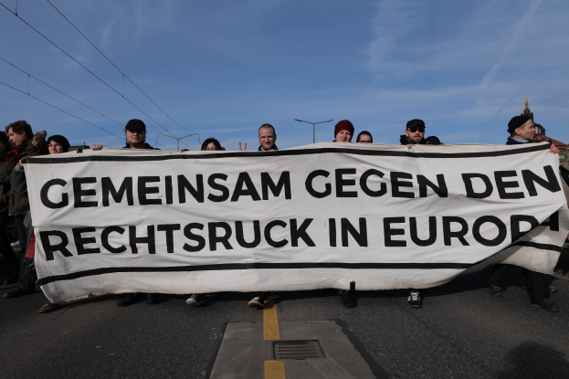 Eine Gruppe von Menschen steht auf der Straße und hält ein Transparent mit der Aufschrift "Gemeinsam Gegen den Rechtsruck in Europa" mit Straßenlaternen, Strommasten, Kabeln, Gebäuden und einem bewölkten Himmel im Hintergrund.