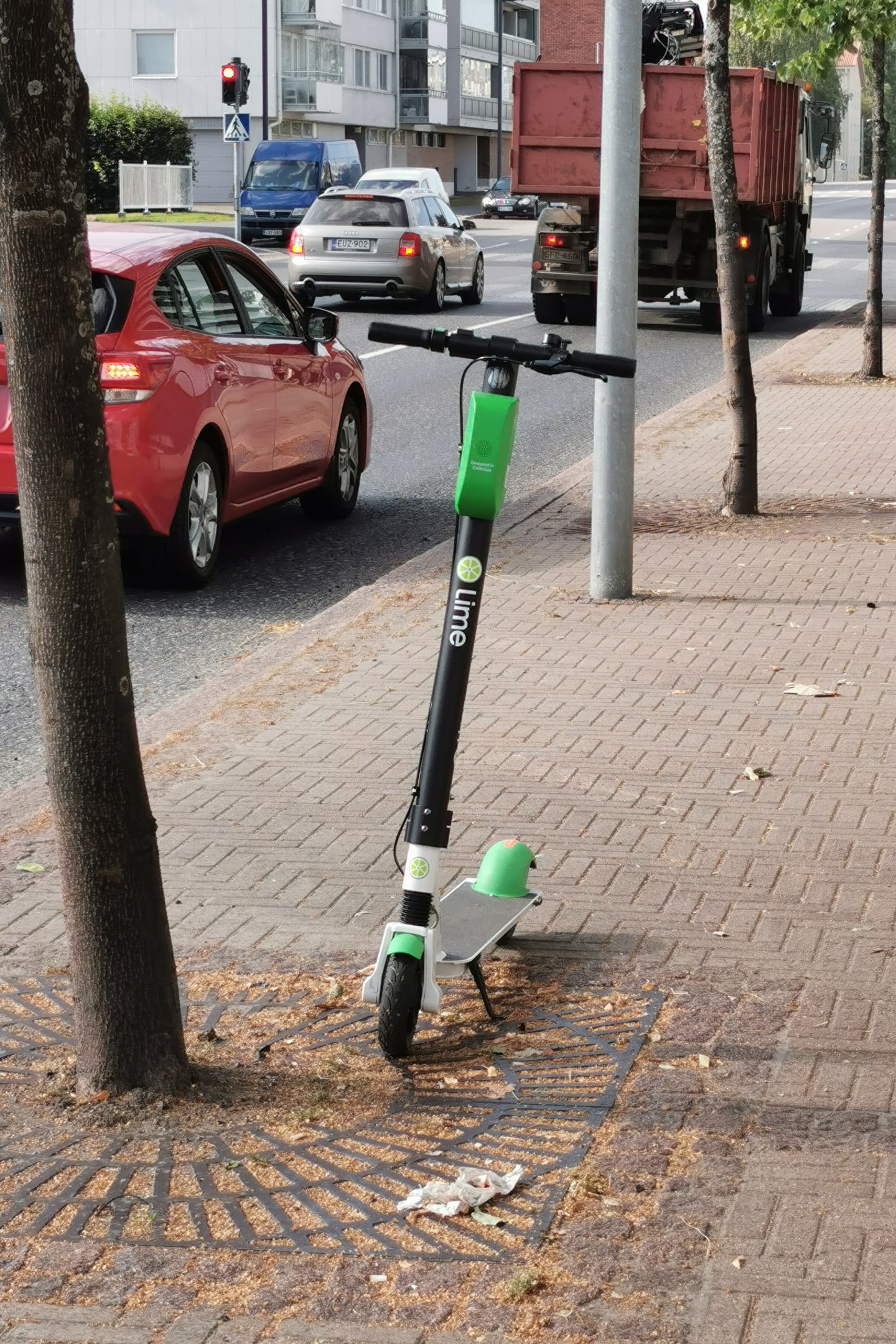 Elektroroller auf dem Bürgersteig neben einem Baum geparkt, mit Fahrzeugen auf der Straße und Gebäuden im Hintergrund.
