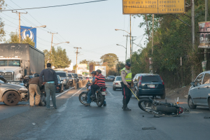Gruppe von Menschen um ein verungl%C3%BCcktes Motorrad am Stra%C3%9Fenrand mit mehreren Fahrzeugen, darunter ein Lastwagen, im Hintergrund und B%C3%A4umen, Masten, Lichter, Schilder und Himmel.
