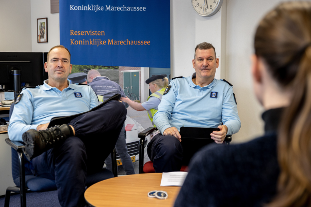 Zwei Polizeibeamte sitzen an einem Tisch mit einer Frau, umgeben von Papieren und Gegenständen, mit einer Fahne, Uhr und Foto Rahmen an der Wand und einem weiteren Tisch mit einem Monitor und Zubehör links daneben.