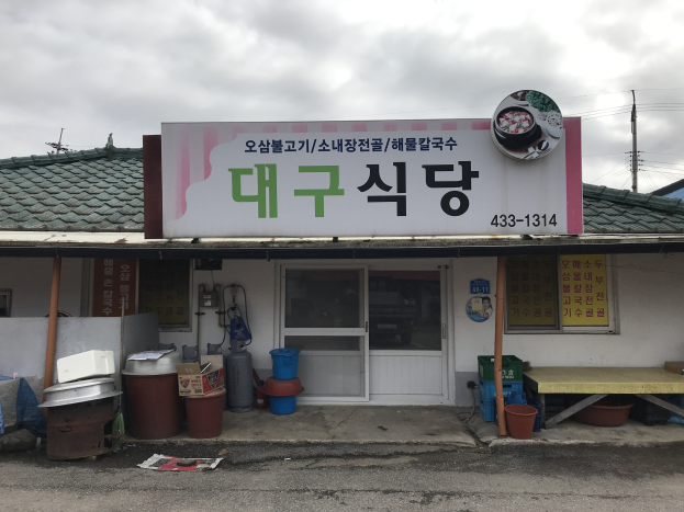 Kleines Gebäude mit einer Tafel, die "Koreanisches Restaurant" lautet, einer Tür, Fenstern, einem Dach und einem bewölkten Himmel.