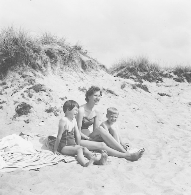 Schwarzes und weißes Foto einer Frau in einem Bikini und zwei Kindern in Badebekleidung auf einer Decke am Strand mit Sand, Gras und Himmel im Hintergrund.