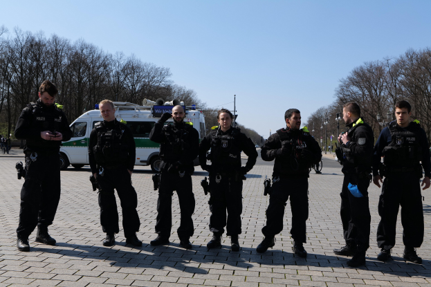 Eine Gruppe von Polizisten steht vor einem Rettungswagen auf einer Straße, die von Bäumen und Laternenmasten umgeben ist und unter einem klaren Himmel liegt.
