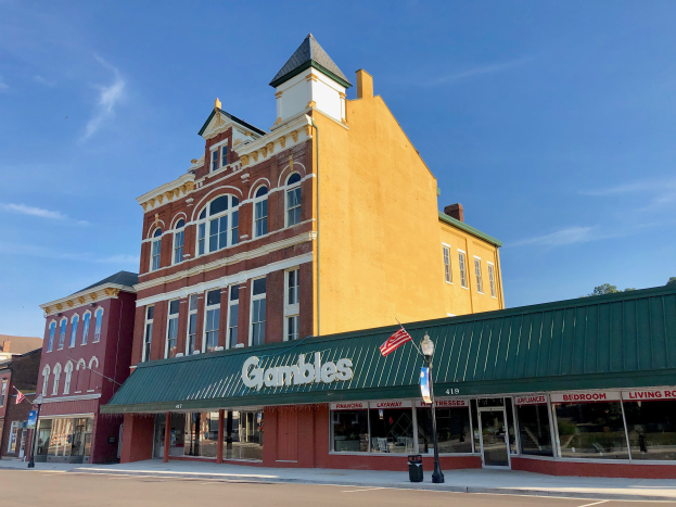 Das Gambles Hotel, ein großes gelbes Gebäude mit einem Uhrenturm, umgeben von Straßenlaternen, Straßenlaternen, Namensschildern, Flaggen, Bäumen und einem bewölkten Himmel.